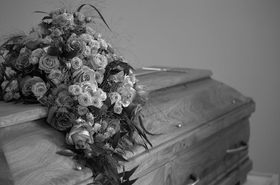 A large, fresh floral arrangement composed of roses in various stages of bloom, predominantly in light and dark shades, resting atop a wooden casket. The bouquet includes a mix of fully opened roses a