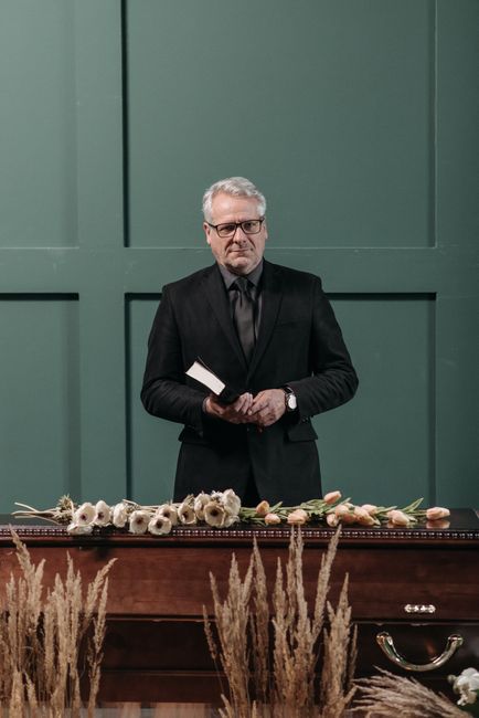 A mature man with gray hair and glasses, dressed in a black suit and tie, stands behind a wooden funeral cask. In his hands, he holds a small book or booklet, and he is positioned in front of a green 