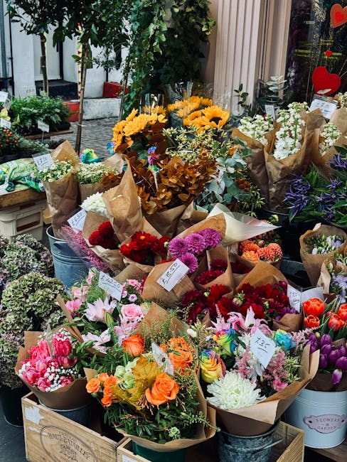 An outdoor display of numerous floral bouquets arranged on a wooden crate and metal buckets. The bouquets feature a variety of fresh flowers, including bright yellow sunflowers, vibrant red and pink r