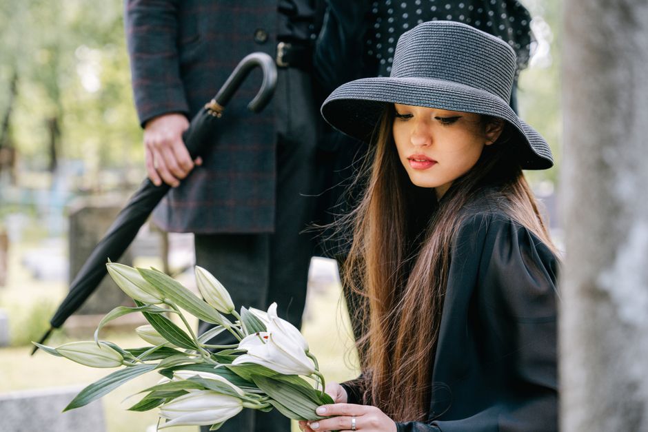 A young woman with long brown hair wearing a wide-brimmed black hat and dark coat is holding a bouquet of white lilies with green leaves, wrapped in clear cellophane. She is standing outdoors in a cem
