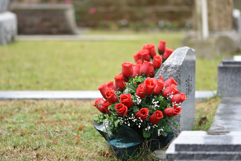 A bouquet of fresh red roses, tightly arranged with green foliage and delicate white baby's breath accents, rests against a grey granite headstone at West Norwood Cemetery. The roses are in full bloom