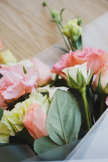 A close-up of a floral arrangement featuring pale pink and creamy white eustoma (lisianthus) blooms with ruffled petals, accented by fresh green leaves and stems. The flowers are arranged in a bouquet
