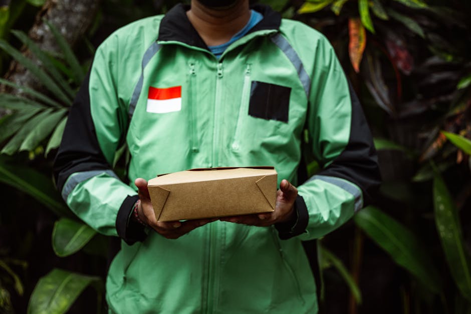 A delivery person wearing a bright green jacket with black and grey accents, featuring a red and white flag patch on the left side of the chest, holds a plain brown cardboard parcel in both hands. The